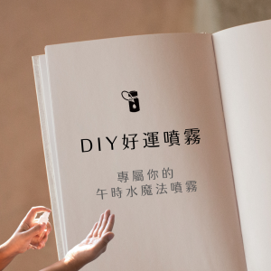 畫面以奶色系精裝書籍空白頁為背景，頁面中央黑色精油瓶圖示下方文字「DIY好運噴霧／專屬你的午時水魔法噴霧」，左下角有雙手正在噴灑精油噴霧，背景柔和窗光斜射在淺米色亞麻布上，整體營造出手作精油噴霧帶來好運與魔法感的氛圍。