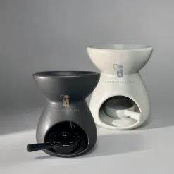 這張圖片展示了兩款 SCENTFANATIVY 品牌的陶瓷精油燈──一款霧面黑色與一款純淨白色。產品採用現代極簡風設計，碗狀上部用於放置香薰蠟或精油，下方圓弧開口可放置茶燭作為加熱來源。高質感陶瓷材質與柔和光影相映，完美呈現產品的工藝細節與質感。這款黑白雙色的「陶瓷精油燈」非常適合用於家居香氛、辦公室擴香、瑜伽室放鬆或送禮，兼具實用與美感。關鍵詞包括：陶瓷精油燈、香薰擴香、家用香氛、極簡設計、黑色精油爐、白色精油爐、SCENTFANATIVY